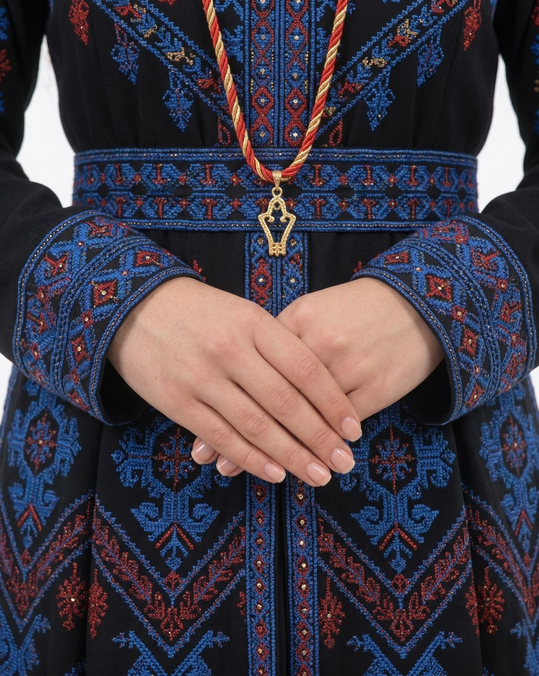 Alhaifawi | Person wearing a black embroidered garment with blue and red patterns, holding hands together.